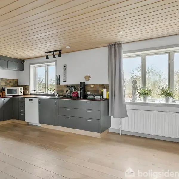 Et moderne køkken med grå skabe og hvidevarer, herunder ovn og mikrobølgeovn, er placeret i hjørnet af et lyst rum med trægulv og et stort vindue med udsigt til træer.