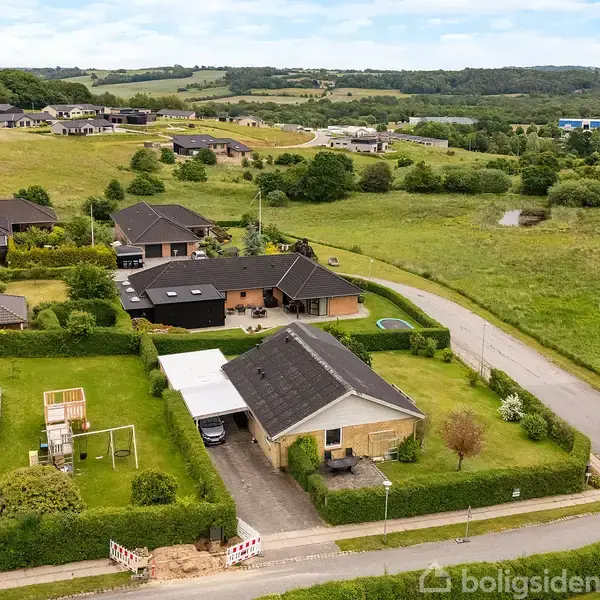 Huse med flade tage ligger på en stille villavej omgivet af grønne marker og små bakker. Et legehus og en indkørsel med carport er synlige i forgrunden.
