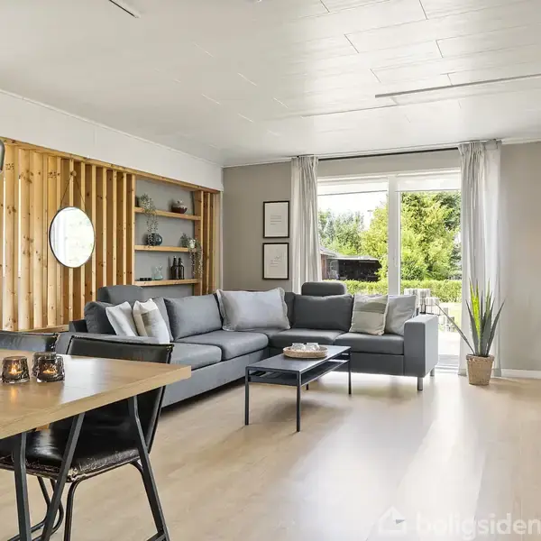 Stue med grå sofa og kaffebord foran en stor åben dør til en terrasse. Ved siden af er der en spiseplads med bord og stole. Indretning med træpaneler og planter.