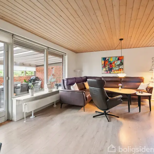 Stue med moderne lædermøbler står ved et sofabord og en farverig maleri på væggen, i et lyst rum med parketgulv og store vinduer, der kigger ud på en overdækket terrasse.