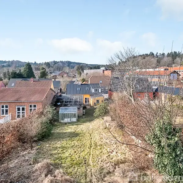 Flere huse med røde tage står stille i en landsby omgivet af buske og græsarealer. I baggrunden ses skovklædte bakker under en blå himmel med lette skyer.