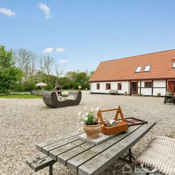 En træbænk står stille på en grusbelagt gårdsplads ved siden af planter i potter. I baggrunden ses et hvidt hus med rødt tag omgivet af grønne træer.