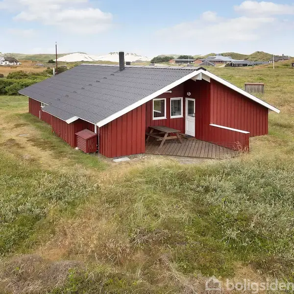 Rødt træhus med sort tag står stille på en grøn græsplæne. Omkring ses andre huse og åbne marker under en blå himmel med skyer.
