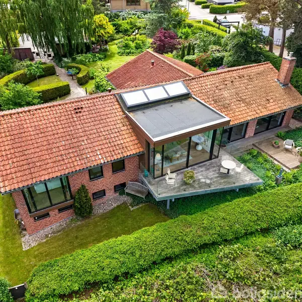 Et stort hus med rødt tag og en stor udestue står i en velplejet have med buske og stier. Udestuen har store vinduer og en terrasse med havemøbler.