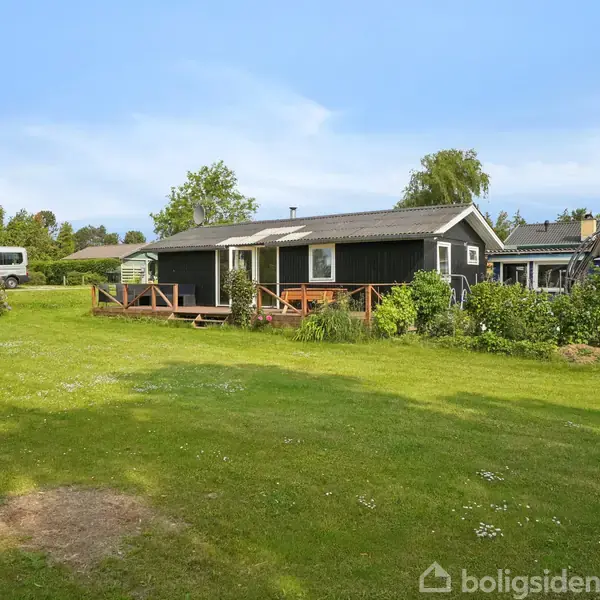 Et sort hus står stille i en frodig have med græsplæne og buske omkring. Flere bygninger og træer ses i baggrunden under en klar blå himmel.
