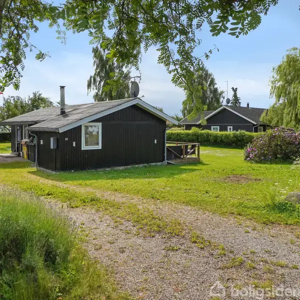 Et sort træhus står på en grusbelagt grund, omgivet af en grøn have med spredte buske og træer. Andre lignende huse kan ses i baggrunden under en blå himmel.