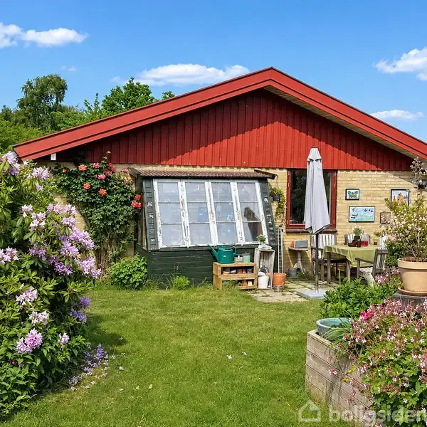 Hus med rød tag og gul mursten omgivet af blomstrende buske. Havebord og parasol står ved siden af huset. Solrig dag med blå himmel og hvide skyer.