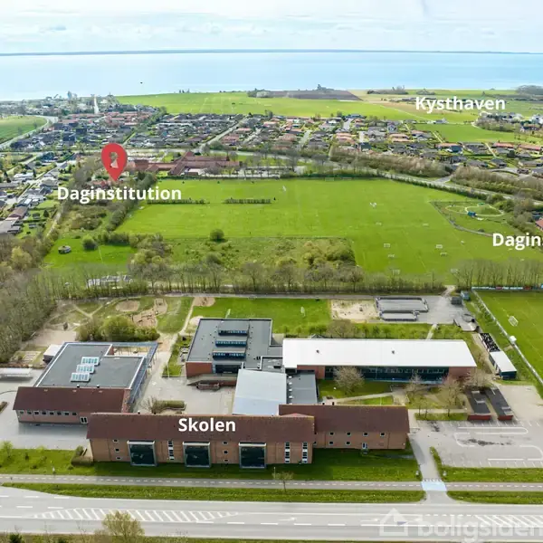 Skolen vises fra luften med boldbaner omkring. Skiltning inkluderer "Skolen," "Daginstitution," og "Kysthaven." Bagved strækker sig grønne områder og en bydel langs kysten.