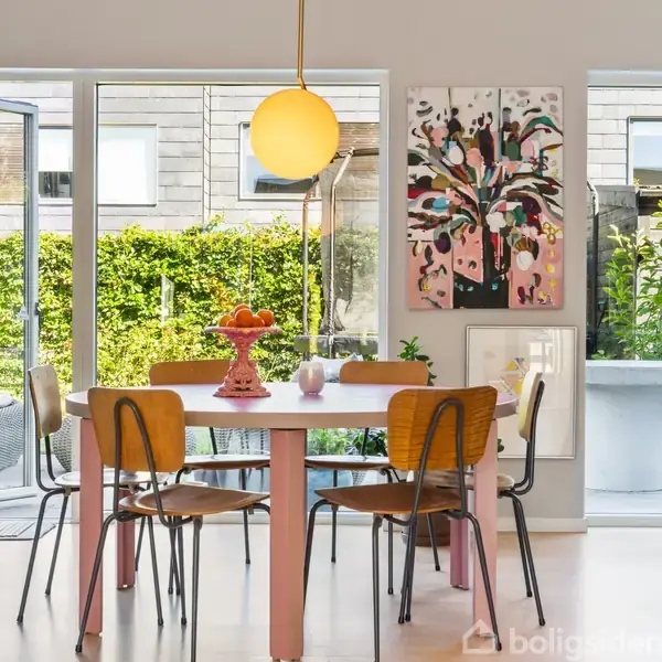 Spisebord med fire stole omgivet af lyse åbne døre, der fører til en have med grønne planter. Rund loftslampe hænger over bordet. Kunstværker pryder væggen.