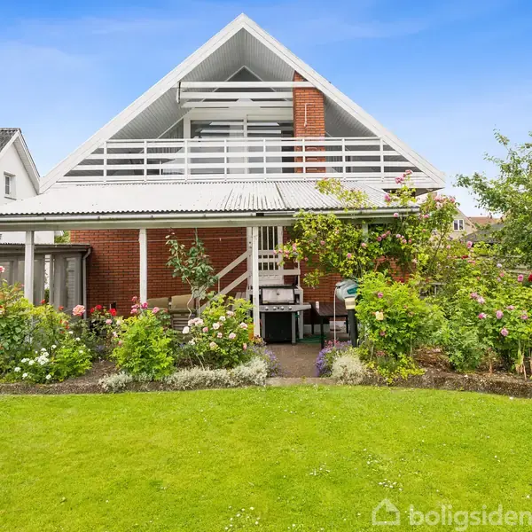 Et moderne hyggeligt hus med A-formet tag i røde mursten og hvide rammer. Huset har en terrasse med trapper. Haven er velholdt med blomster og en stor græsplæne.
