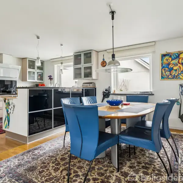 Spisebord med blå stole står på tæppe i et lyst køkken-alrum. Bagved ses sorte skabe, hvide vægge og kunstværk med farverigt motiv.