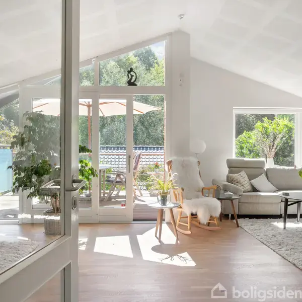 Stue med lyst interiør og store, lyse vinduer, der åbner ud til en balkon med parasol. Komfortabel sofa, lænestol og sofabord omgivet af grønne planter.