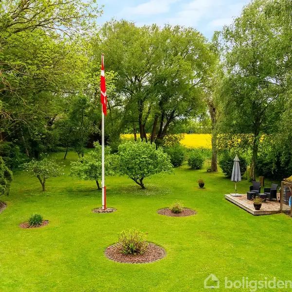 Flagstang med dansk flag står i en grøn have; omgivet af træer, blomsterbede og en terrasse med møbler. I baggrunden ses en mark med gul afgrøde.