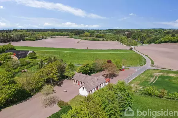 En hvid bygning med rødt tag står stille omgivet af grønne marker og træer. En snoet vej løber gennem det landlige landskab under en blå himmel med spredte skyer.