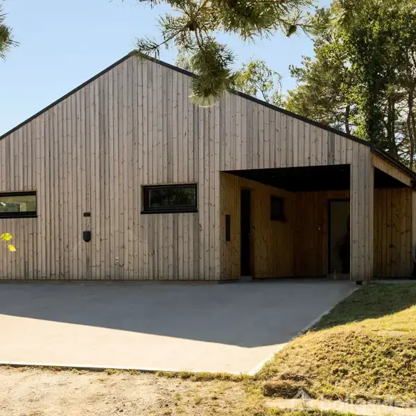 Et moderne træhus med skråt tag står i en skovklædt omgivelse. Foran huset er en garage, og flisebelægning fører op til indgangen.