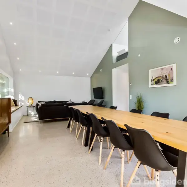Langt spisebord med sorte stole omkring står i en moderne og rummelig spisestue. Til venstre ses en skænk med dekorationer. Væggene er hvide med én grøn væg.