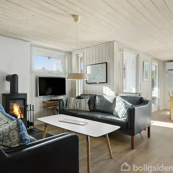 Sort lædersofa står ved siden af en tændt brændeovn i en lys stue. Stuen er forbundet med en spisestue fyldt med naturlig belysning.