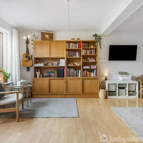 En stue med trægulv, reol fyldt med bøger og pyntegenstande. En guitar hænger ved reolen. Store vinduer til venstre med planter. Fladskærm på væggen og komfortable stole.