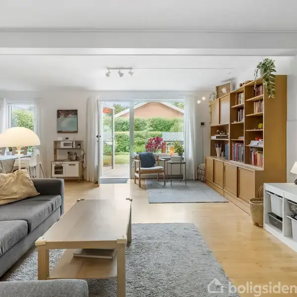 En stue med grå sofa og sofabord på et tæppe. Udendørs lys strømmer ind gennem glasskydedøre, der fører til en have. Reoler fyldt med bøger står mod en væg.