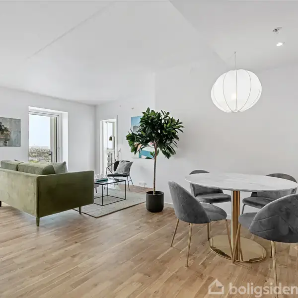 En stue med grøn sofa og trægulv, hvor en rund spisebord med fire grå stole står under en hvid lampe. Store vinduer giver udsigt og lys, og planter pynter rummet.