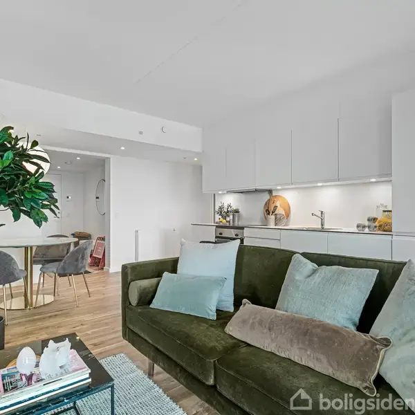 Grøn sofa med puder står i lys stue, ved siden af glasbord med bøger og planter, i et rum med åbent køkken og spiseområde bagved.