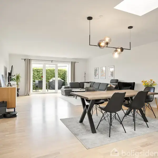 Spisebord med sorte stole står på et tæppe, omgivet af moderne, lys stueindretning med sofa. I baggrunden ses store vinduer med udsigt til en hæk. Ingen synlig tekst.