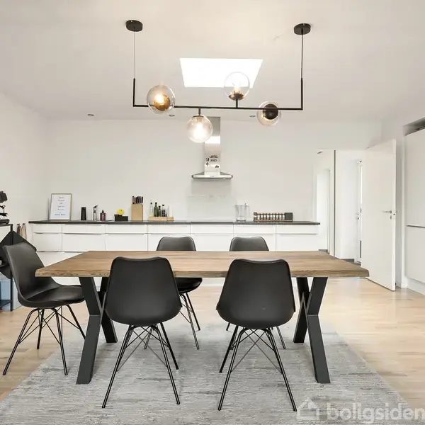 Spisebord med sorte stole står under moderne loftlamper i et lyst køkken-alrum med hvidt køkkenudstyr og træhylder. Til venstre ses en reol med planter og køkkenudstyr.