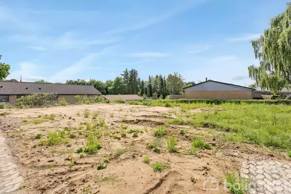 Tomt med ujævn jord og spredte græsstrå ligger mellem to huse i et boligområde. Omkringliggende træer og blå himmel skaber en rolig atmosfære.