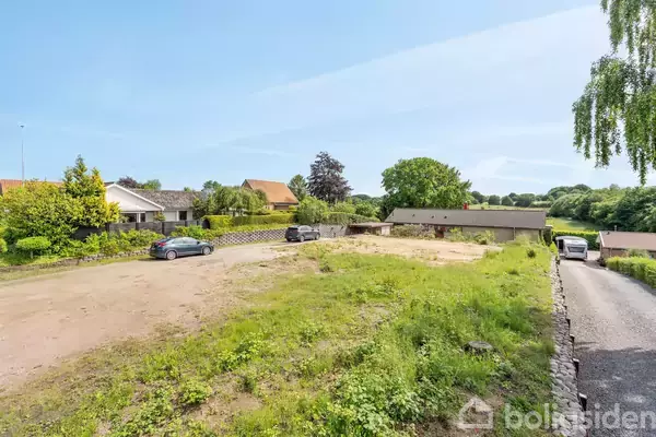 Biler holder parkeret på en grusplads omgivet af grønne områder og huse. En lang, smal vej fører ind til området. I baggrunden ses flere træer.