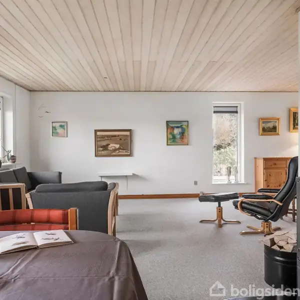 En stue med en brændeovn, der brænder. Til venstre står en reol og sofa ved vinduet. Malerier hænger på væggen, og et spisebord er i forgrunden med en kikkert og bog.