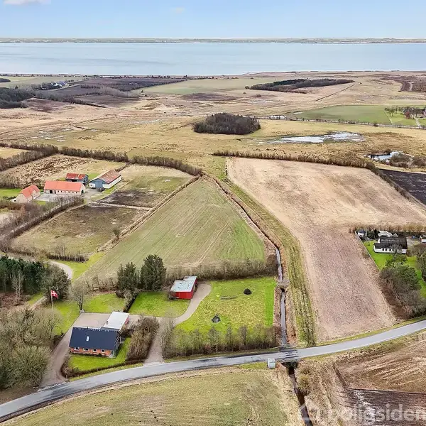 Luftfoto af landligt område med huse og marker. Huse er omgivet af træer og græsarealer. Baggrunden viser en stor sø eller fjord omgivet af marker og spredte bygninger.