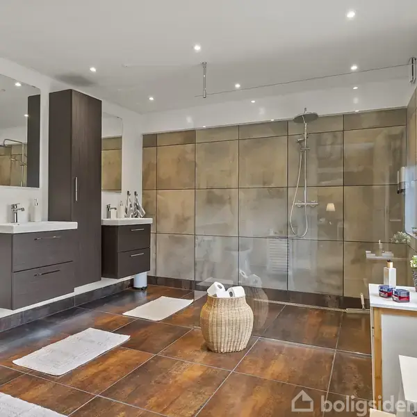 Et rummeligt badeværelse med en bruseniche, mørke træmøbler, og tre håndklæder på en holder. Gulvet er dækket af hvide tæpper, og vinduet har sorte persienner.