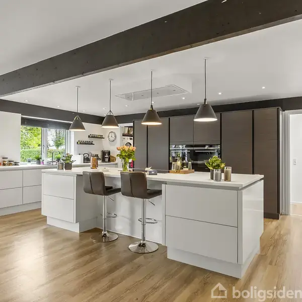 Et moderne køkken med en stor ø-køkken, hvor der står tre barstole. Køkkenet har træ-look gulv, hvide skabe, sorte indbyggede ovne og store vinduer med grøn udsigt.
