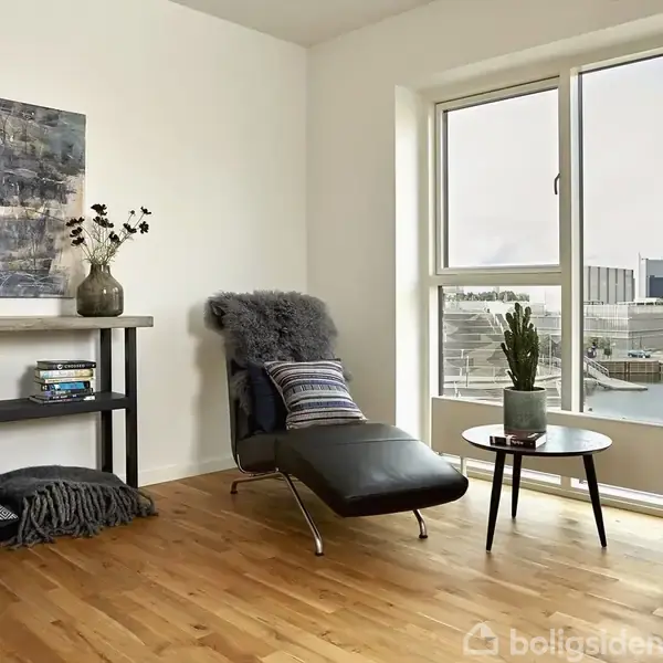 En moderne lænestol med pude og tæppe står ved et stort vindue med udsigt over en by. Et lille rundt bord har en plante. Kunst og dekorative genstande pryder væggen og en hylde.