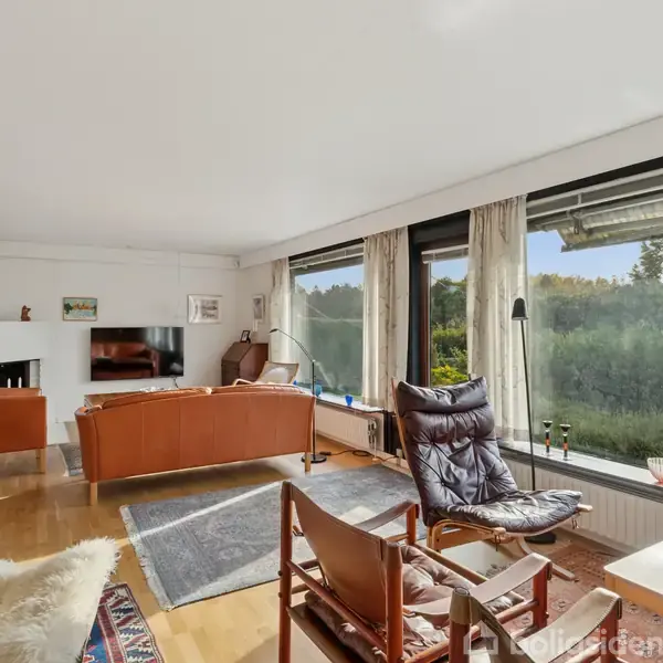 Stue med orange sofaer og lænestole vendt mod en pejs, store vinduer med udsigt til grønne træer. Parketgulv, tæpper og små borde bidrager til den hyggelige atmosfære.