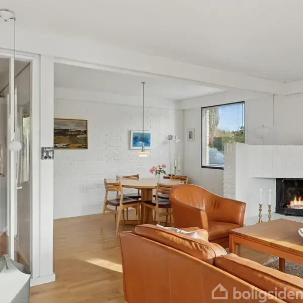 En stue med brune lædersofaer foran en tændt pejs. Et rundt spisebord står i baggrunden ved siden af et vindue, omgivet af lyse vægge og billeder.