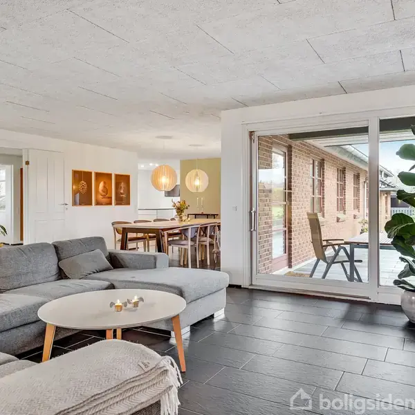 En grå sofa står i en moderne stue med et rundt sofabord foran. Gennem glasskydedøre ses en terrasse med stole, og spisestuen er i baggrunden med træmøbler.