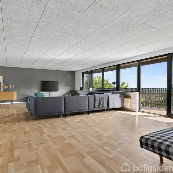En stue med en blå sofa i et åbent rum, hvor sofaen er rettet mod en fladskærm. Store vinduer giver udsigt til en balkon og natur udenfor.