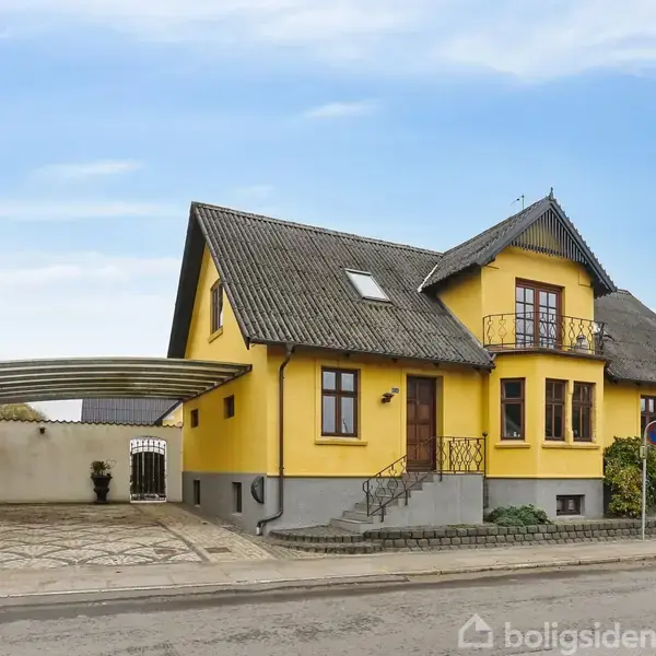 Gul to-etagers hus med grå tagsten står ved en vej. Det har en lille trappe ved hoveddøren og en carport på venstre side, omgivet af andre huse.
