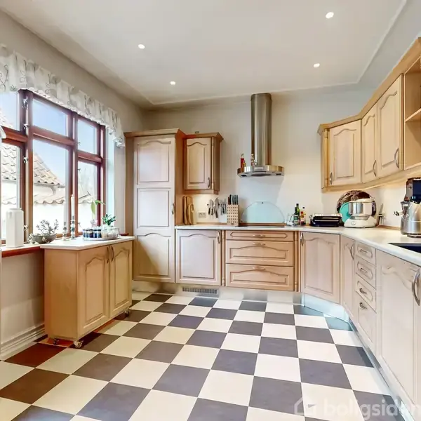 Køkken med træskabe og -skuffer er ryddet. Det har en indbygget ovn, emhætte, og diverse køkkenredskaber. Brune og hvide tern på gulvet. Store vinduer giver naturligt lys.