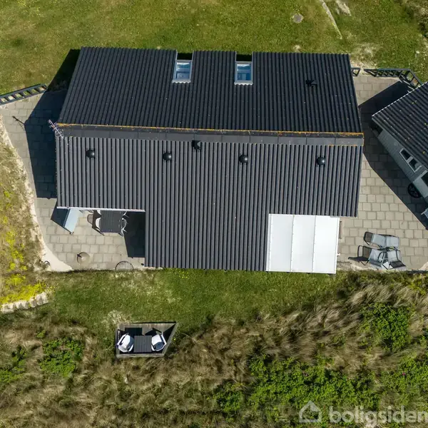 Sort taghus med terrasse set fra luften. Taget har flere ovenlysvinduer. Huset er omgivet af grønne områder og flisebelagt terrasse med havemøbler.