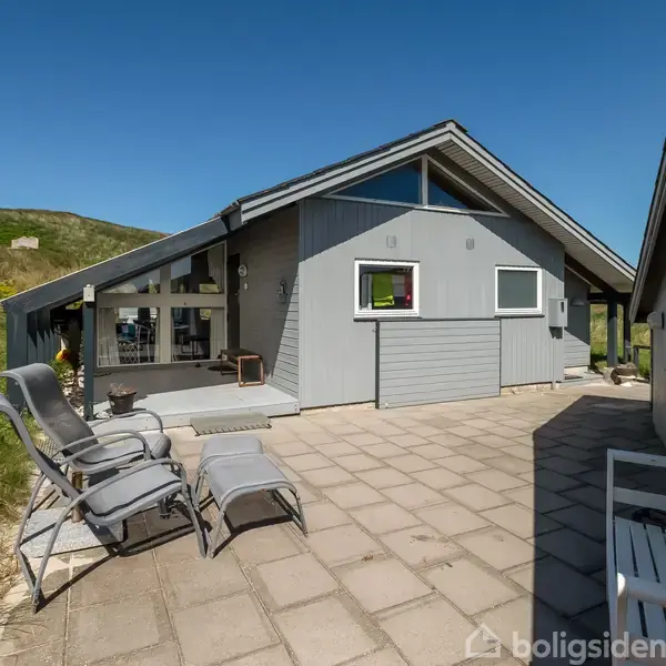 Et gråt sommerhus med store vinduespartier står på en flisebelagt terrasse med liggestole, omkranset af en grøn bakke og klar, blå himmel.