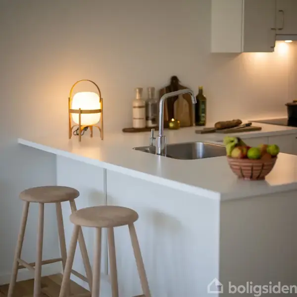 En moderne køkkenø med to træskamler står i fokus. En skål med frugt og en tændt lampe ligger ved siden af en vask. Baggrunden har krydderier og en skærebræt.