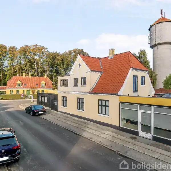 En gade med parkerede biler foran en hjørnebygning med rødt tag. I baggrunden står et vandtårn og flere huse omgivet af træer.