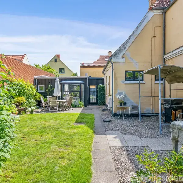 En gul villa står i en velholdt have med græsplæne og grussti. Havemøbler er placeret under en parasol, og en lille buegang fører til flere bygninger i baggrunden.