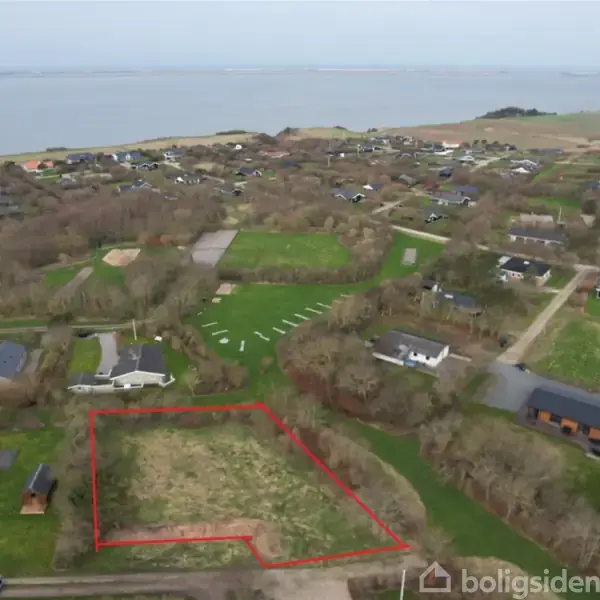 Grundstykke markeret med rød linje ligger i et villakvarter nær vandet. Omkringliggende huse er placeret blandt træer og grønne områder tæt ved kysten.