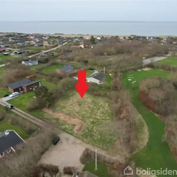 Rødt pilmarkeret stykke jord ligger centralt blandt spredte huse, tæt på en vej. Området er grønt og åbner op mod havet i baggrunden.