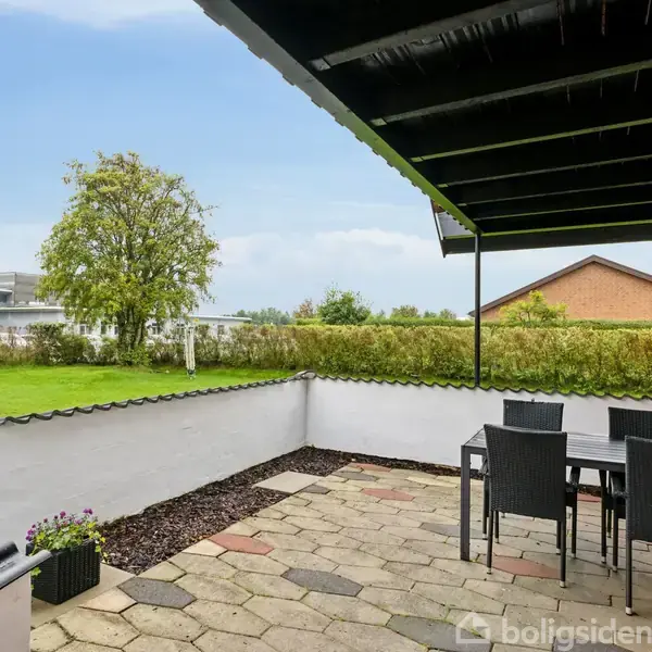 Terrassemøbler står på flisebelagt terrasse under halvtag. I baggrunden ses en velplejet græsplæne med buske og træer, omgivet af en hvid mur. En rød murstensbygning ses til højre.