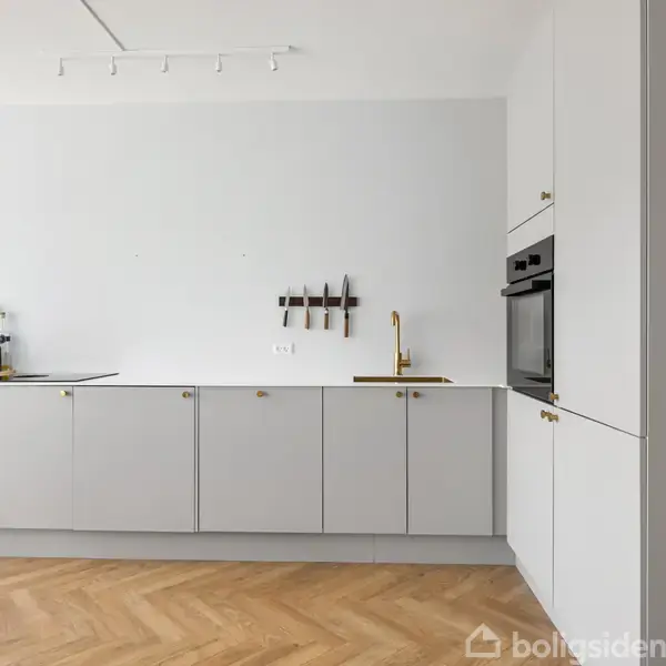 Lysegråt køkken med minimalistiske skabe, indbygget ovn og komfur. På væggen hænger knive ved siden af gyldne vandhaner. Parketgulv og hvid væg supplerer det rene design.
