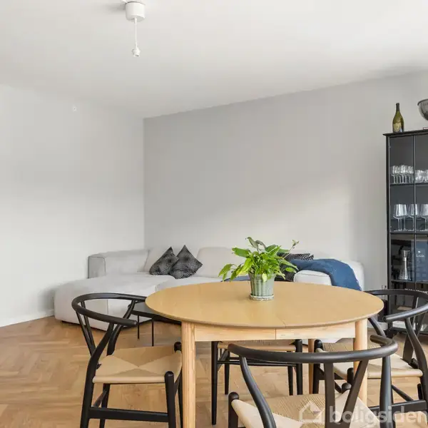 Et rundt spisebord med stole står centralt i en stue med parketgulv. En plante er placeret på bordet. I baggrunden ses en grå sofa og et vitrineskab.